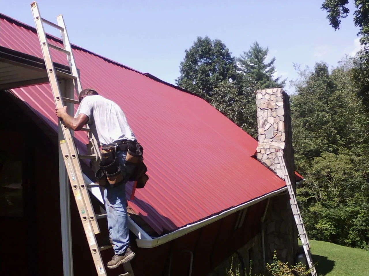 Roofer on a ladder beside a red metal roof and stone chimney — Soffit & Fascia Repair by Roofing Brothers in Moville
